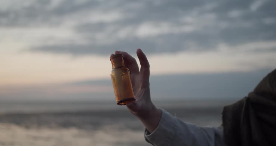 A woman holds a small bottle with a message in it in her hand near the ocean at sunrise