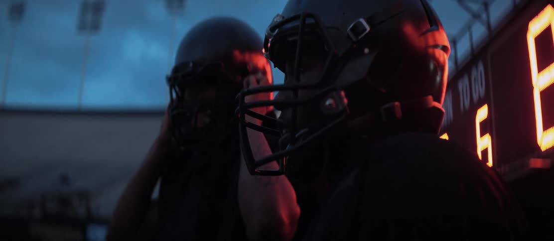 Two teenage football players stand and adjust their helmets in front of a scoreboard in an empty stadium