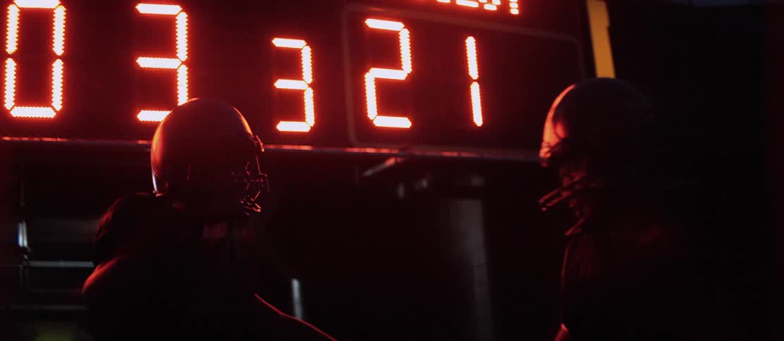 Two men with football helmets and gloves celebrating with a scoreboard behind them