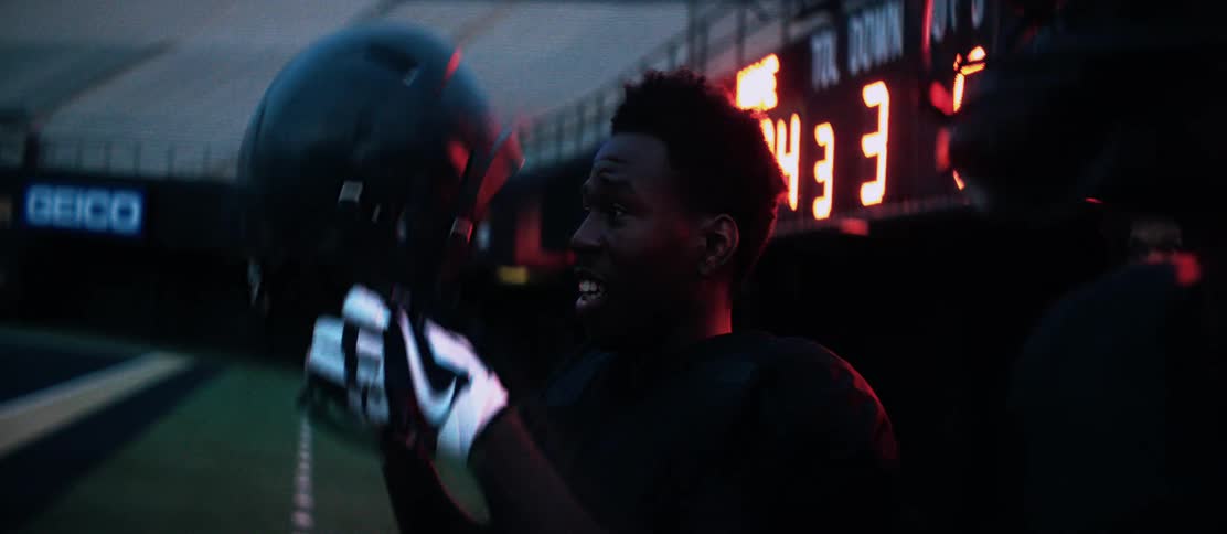 A black football player takes off his helmet as he stands with teammates on the sideline of a football field