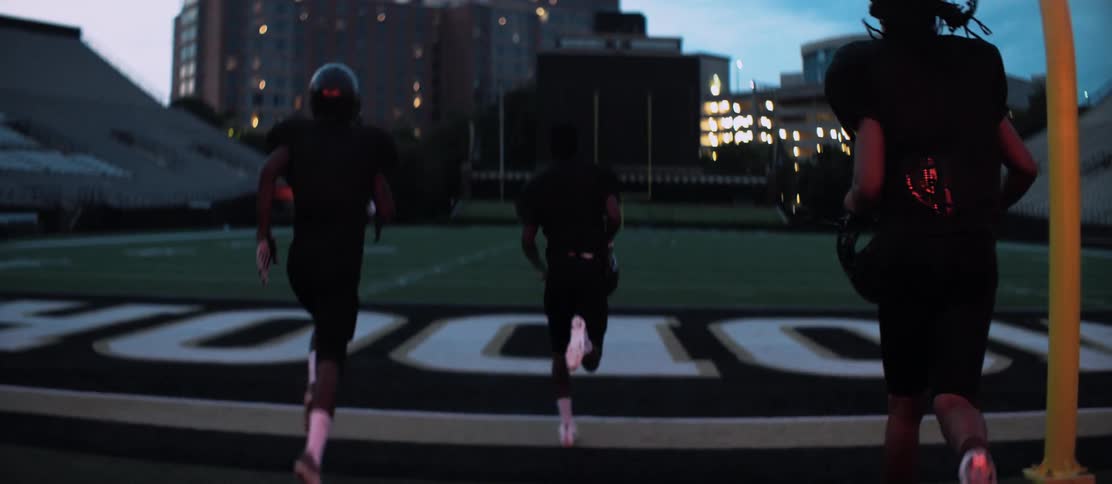 Three football players in uniform run onto the football field at night