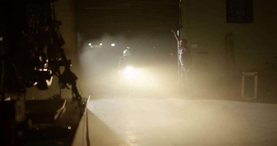A white man watches another man drive a motorcycle into a garage at night