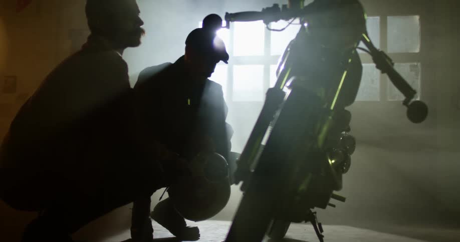 Two men crouching down by a motorcycle is silhouetted against the sunlight coming through windows