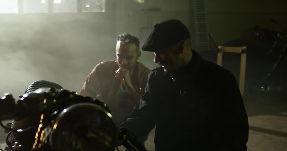 Two white men stand next to a motorcycle in a garage and talk