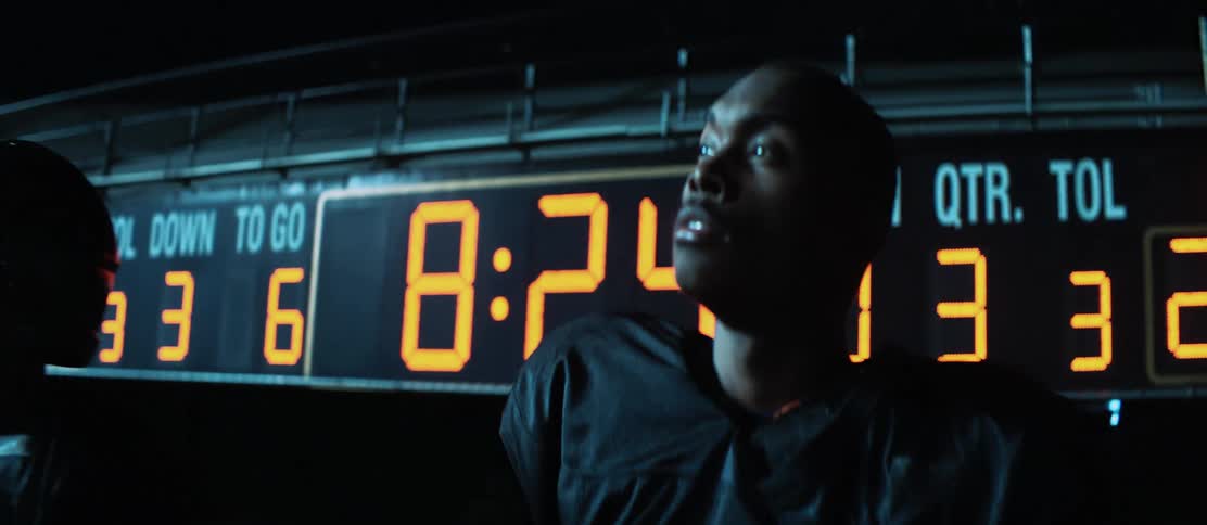 Three football players stand in front of the scoreboard at night