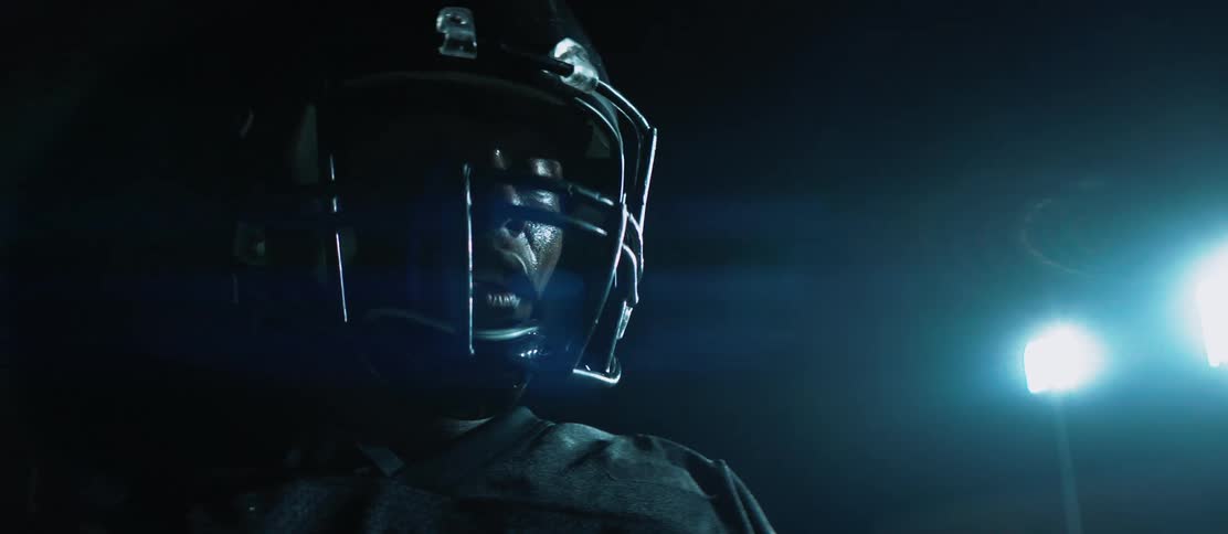 A man with a football helmet and gloves wiping sweat off his face with lights in the background