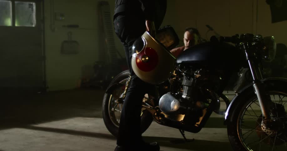 A man kneels in a garage and looks at a motorcycle as another person stands next to it holding a helmet