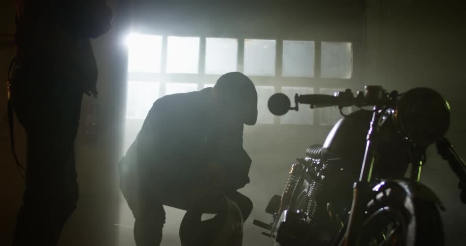 One man stands by as another man bends down to look at a motorcycle