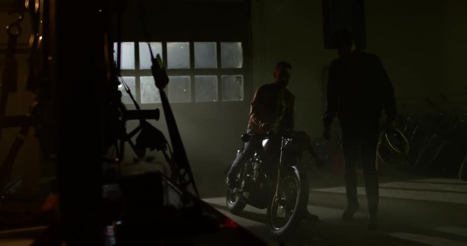 Two men look over a motorcycle in a garage with another motorcycle strapped down to a table