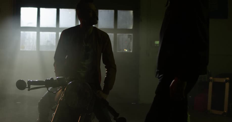 A white man on a motorcycle talks to another man in a garage