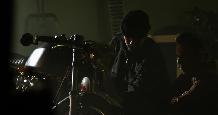 Two white men sit next to a motorcycle