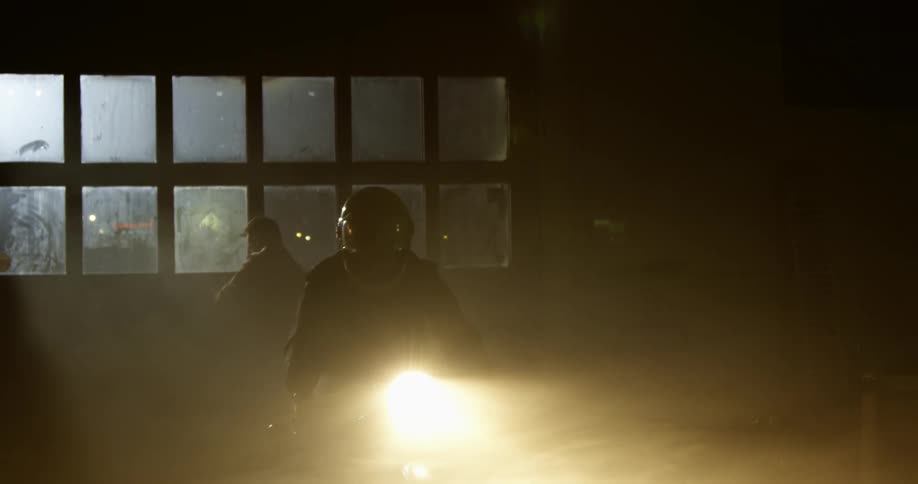 Male rides motorcycle with headlight on into the bay of the garage as another male closes the door behind him