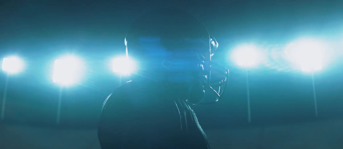 A person in a football jersey and helmet stands in an empty stadium under the bright lights that surround the field