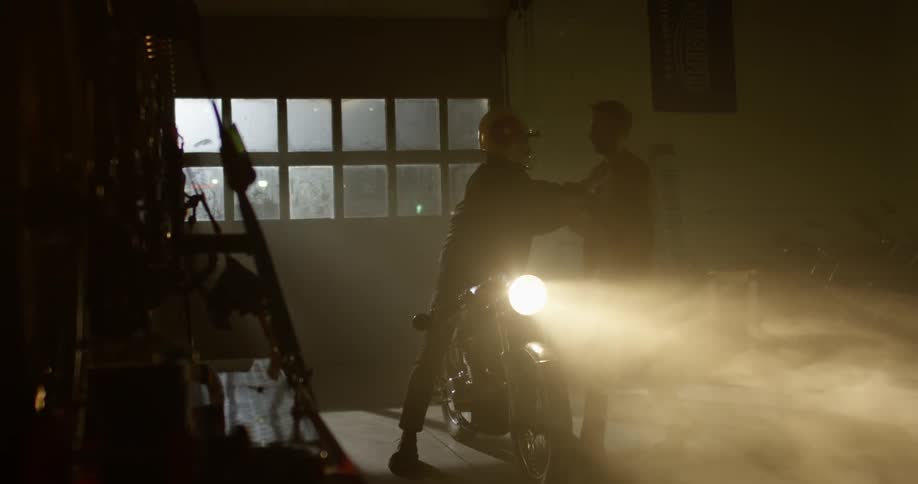 A man sits on a motorcycle and another man walks up to him and they shake hands and hug