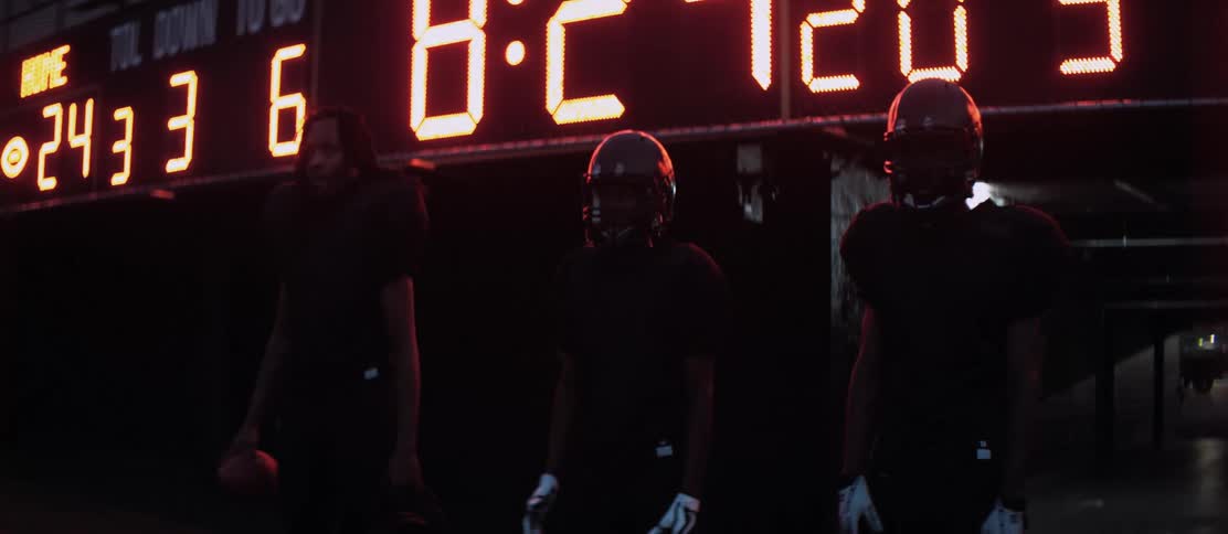 Three black football players stand in front of a scoreboard and stare across the field