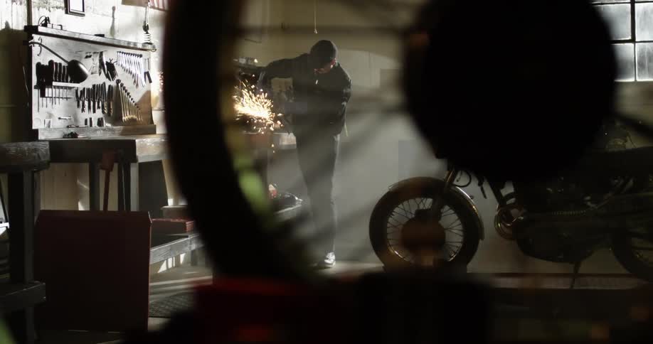 One man welds in a shop as another man looks at a motorcycle