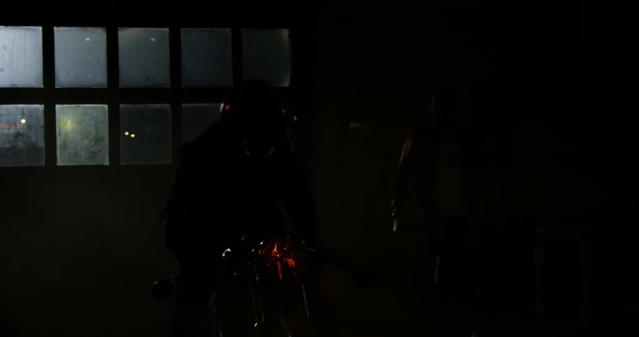 A person sits on a motorcycle inside a dark garage and turns off the headlight