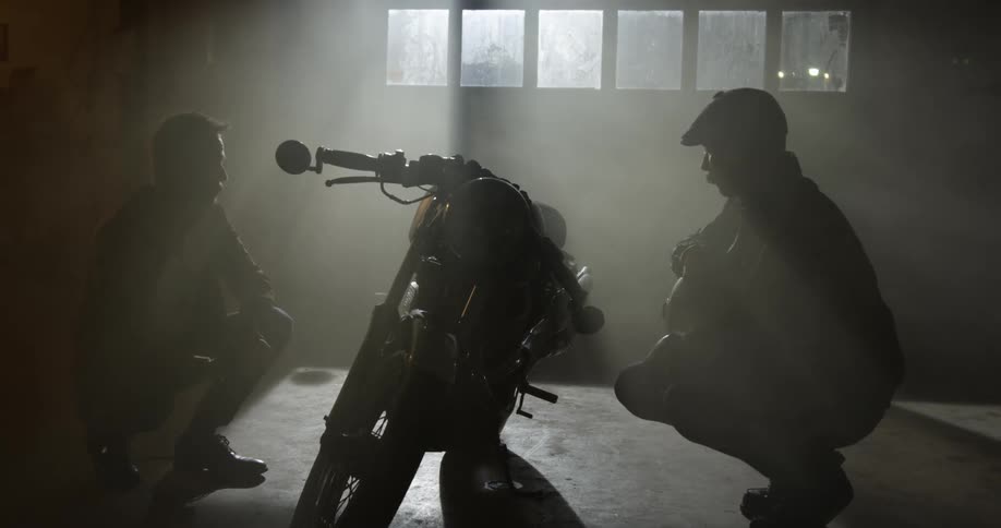 Two men crouching down by a motorcycle is silhouetted against the sunlight coming through windows at the garage