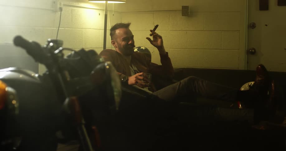 A man with a cigar laughs while sitting on a sofa near a motorcycle in a garage