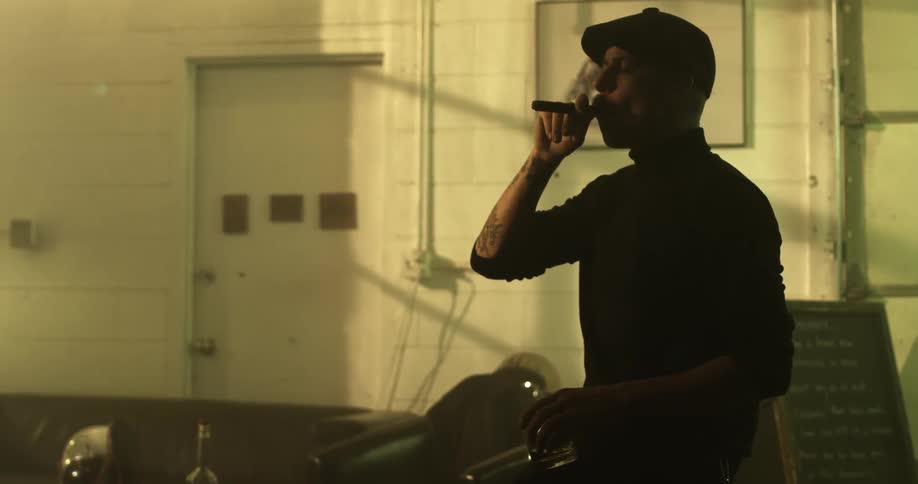 Man with a black beret and asweater holds a glass and smokes a cigar