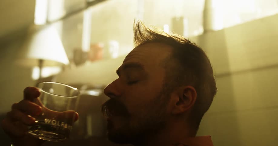 A white man drinks liquor from a glass while sitting indoors