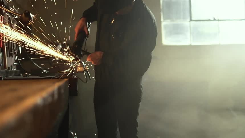 A white man works with tools at a bench that kick off a stream of sparks