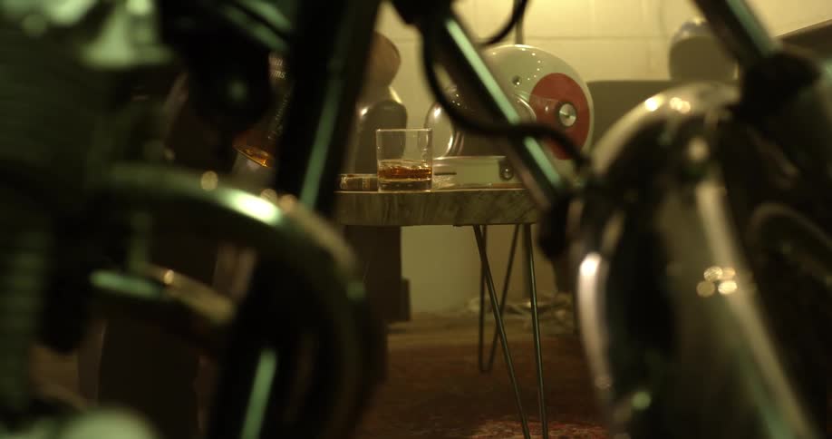 A man pours a drink of alcohol on a table with a motorcycle standing nearby