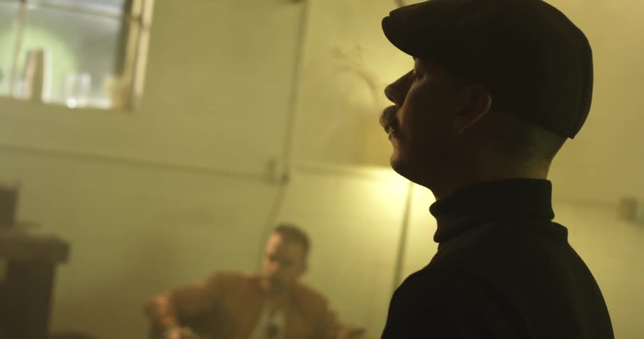 Man smokes a cigar as another man sits nearby smoking one also