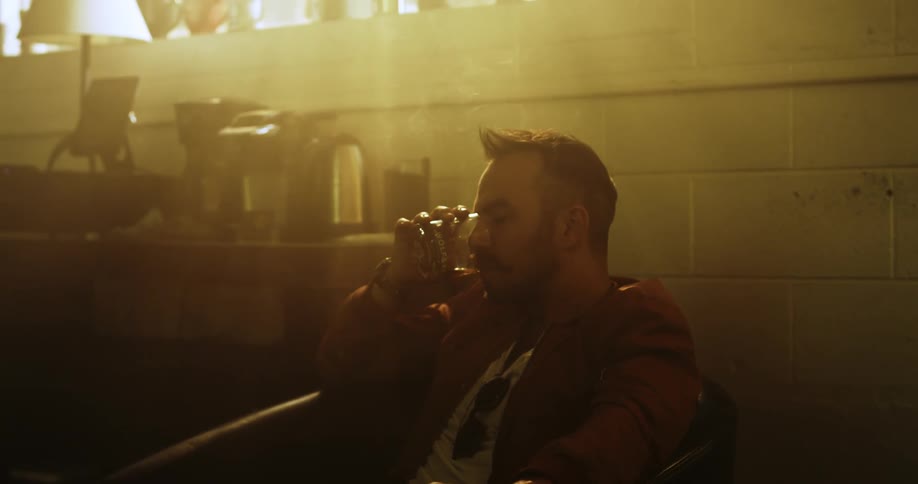 A bored looking white man drinks and smokes while sitting in a chair