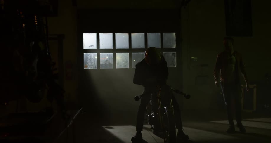 A man walks towards another man sitting on a motorcycle in a garage