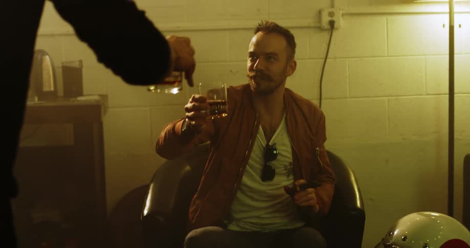 A white man sits in a chair with a cigar and toasts his friend with a drink