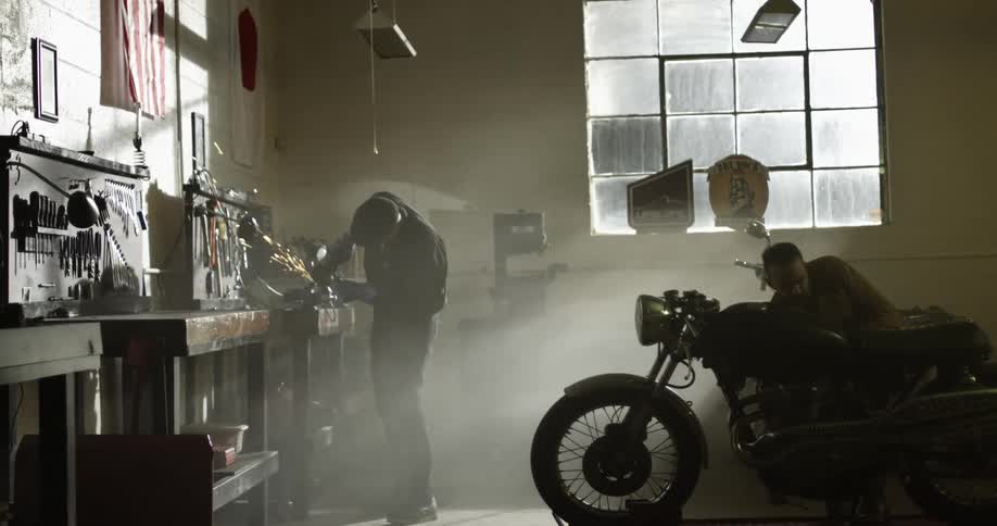 One man in a shop welds while another man looks at a motorcycle