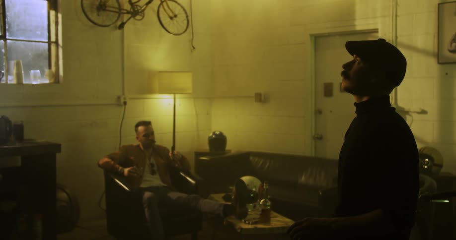 Two white men hang out in a cluttered area indoors