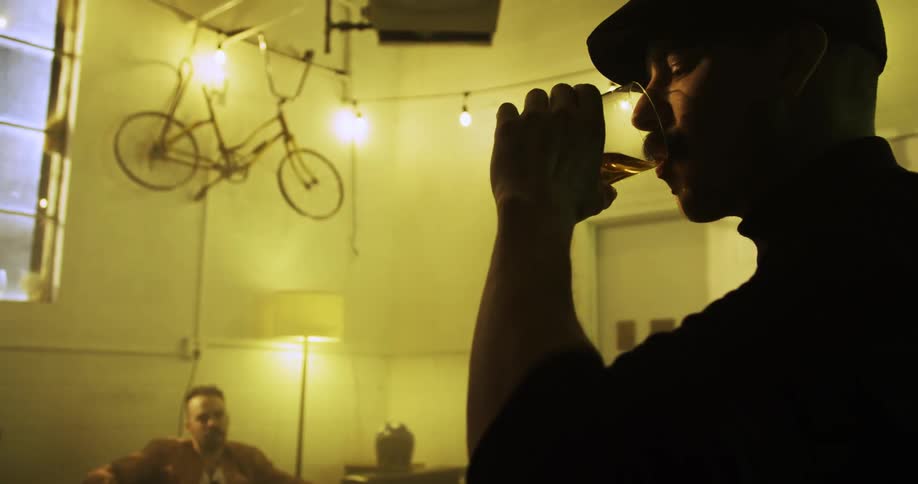 Two men drink and one smokes in living room with bicycle hanging high on wall