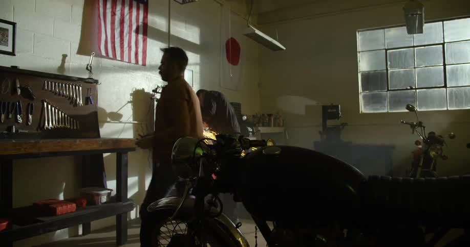 A white man walks through a garage where another man works on a workbench