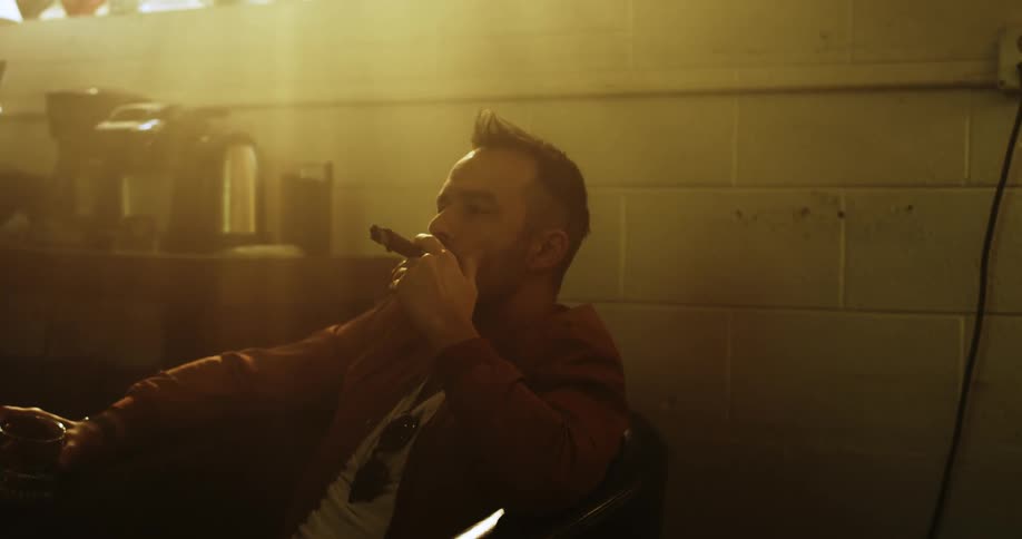 A man smokes a cigar while holding a mug and sitting in a chair