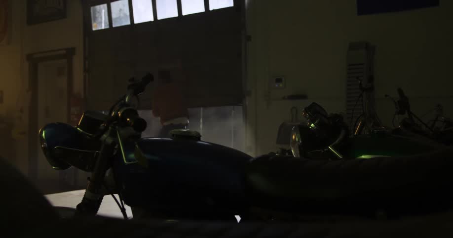 Male rides motorcycle into the garage of a repair shop as the mechanic closes the garage door and the sunlight beams through the door windows