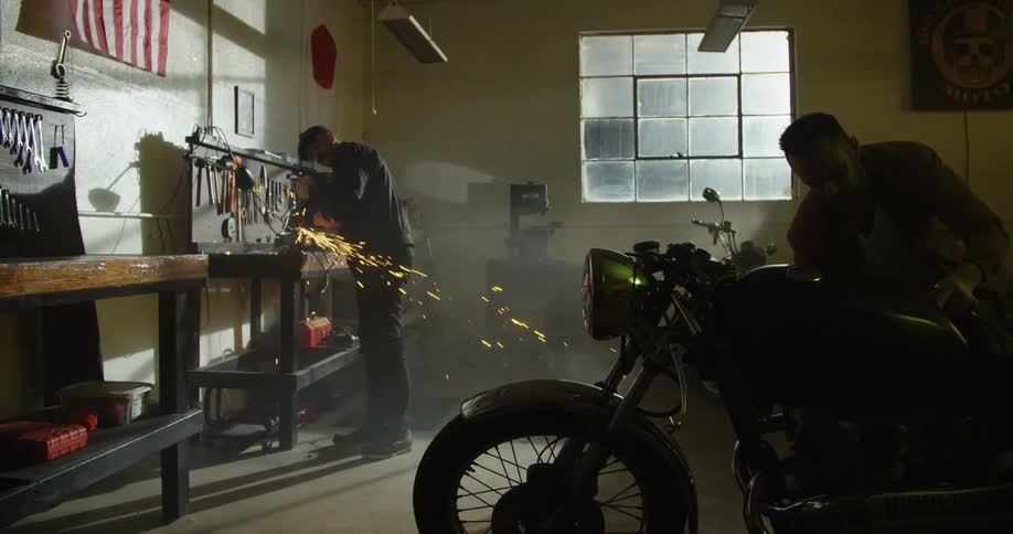 A man in a garage using an electric grinder with sparks while another man works on a motorcycle
