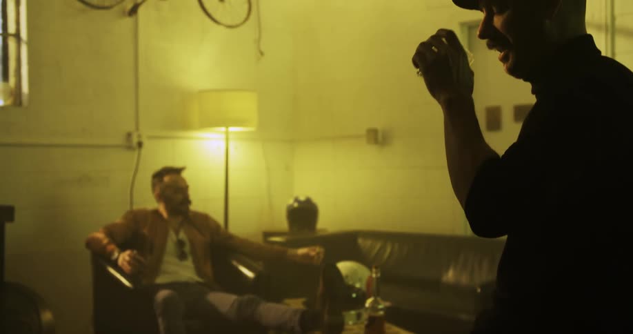 Two white men sit in a living room and drink and smoke