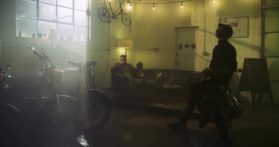 Two white men sit around in a garage smoking and drinking