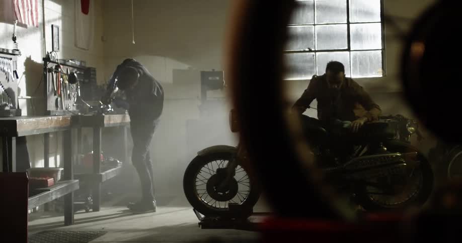 One man welds in a garage as another man looks at a motorcycle