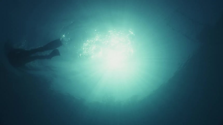 A swimmer in flippers swims out of frame allowing the sunlight to shine through the surface of the water and into the camera