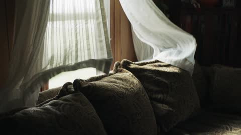 A couch with a open window with curtains blowing in the wind behind it