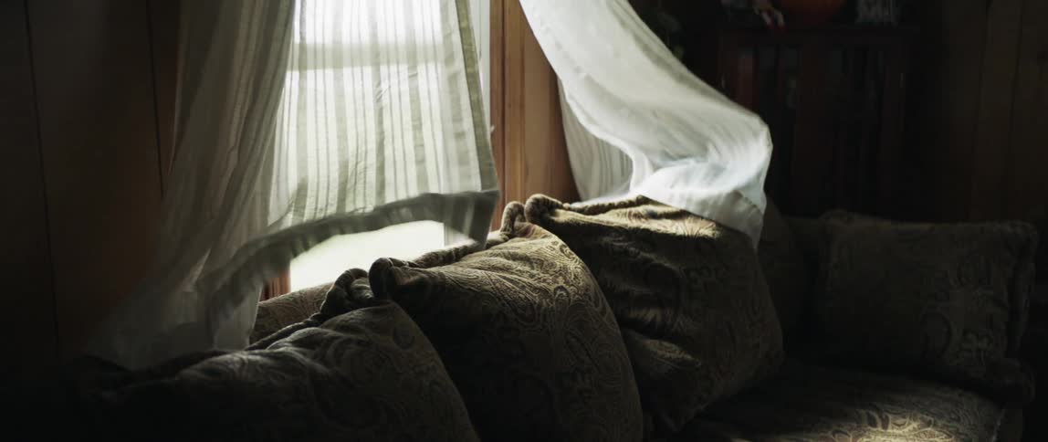 A couch with a open window with curtains blowing in the wind behind it