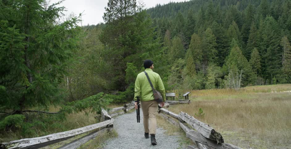 A white male carrying a tripod walks down a gravel trail in a forest