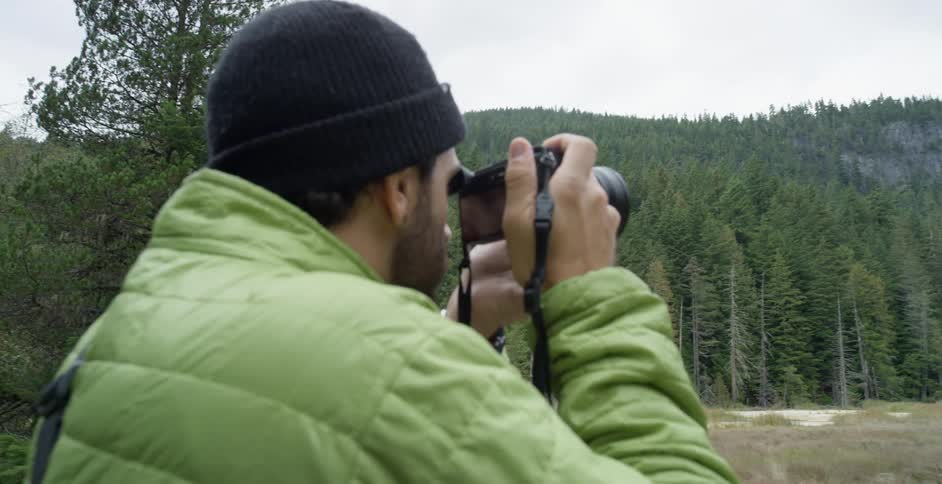 Male photographer with a black beanie and green coat photographs mountain forest