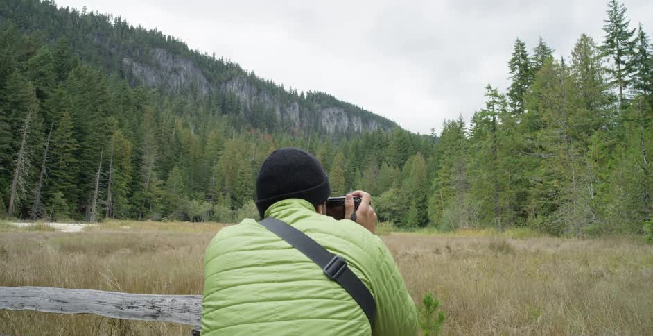Male photographer with a black beanie and a green coat photographs trees in the forest
