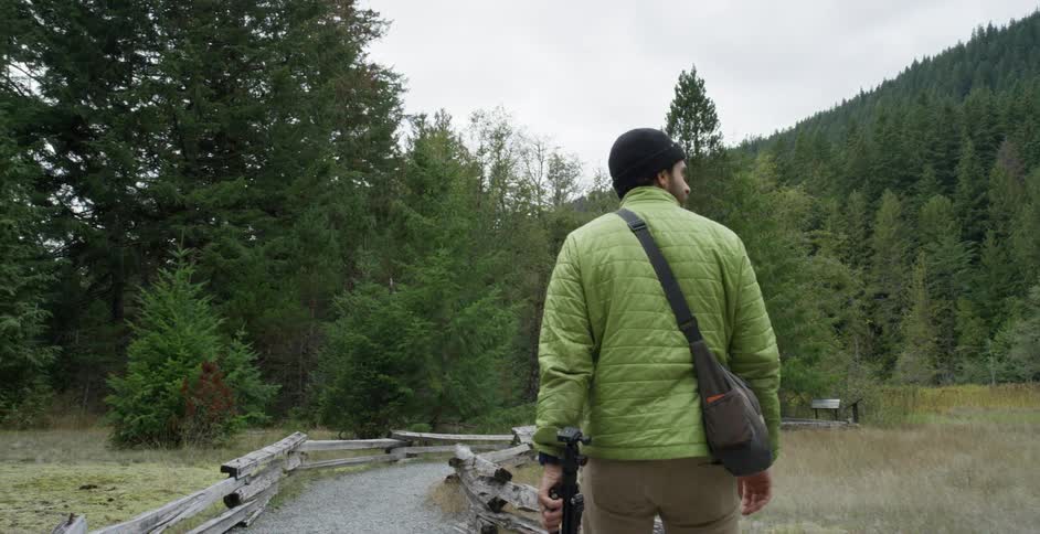 A white man toting a bag and tripod walks on a path near a forest