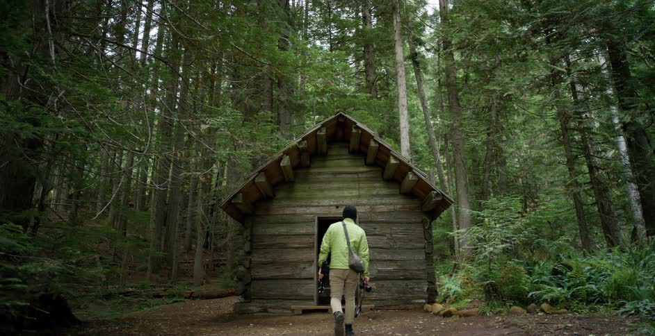 A white man in a coat and hat carries a camera and tripod to a cabin in the woods