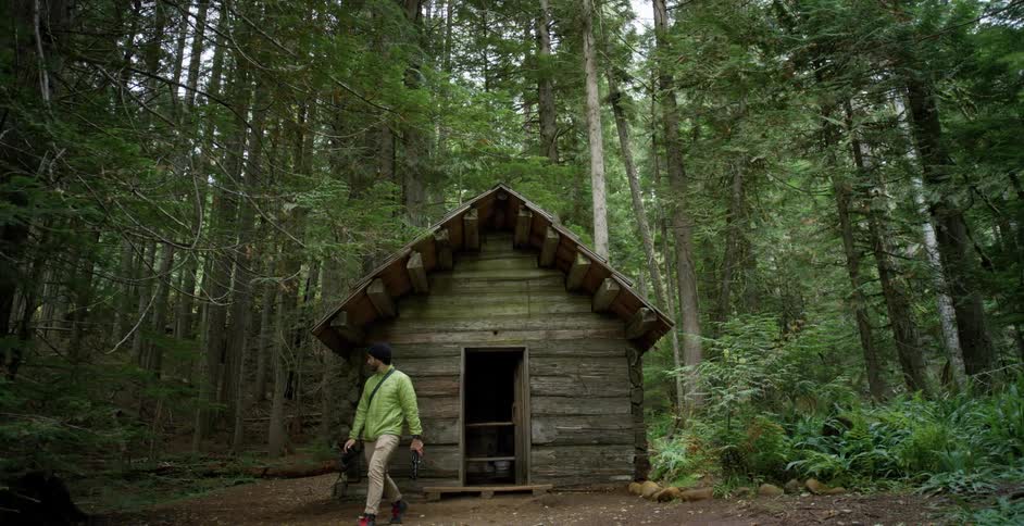 Male steps out of the log cabin in the middle of a wooded area carrying a camera and tripod and continues walking